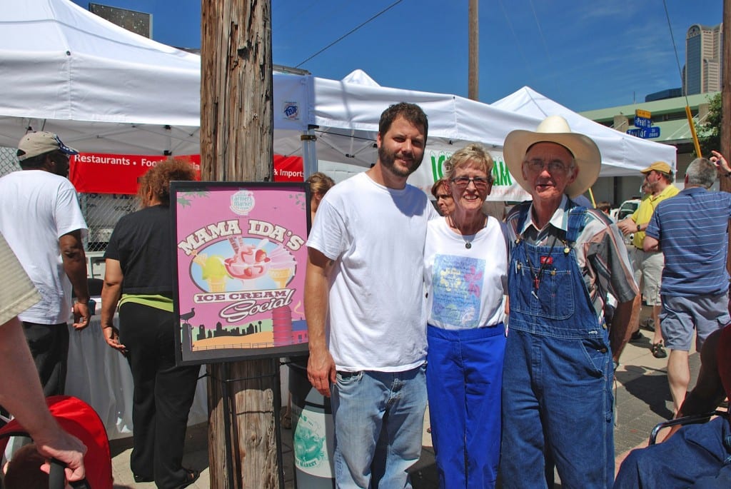 Ida 'Mama Ida' Papert at the Mama Ida's Ice Cream Social event at Dallas Farmers Market, with a young man on her left and an older gentleman in overalls on her right. The event sign behind them reads 'Mama Ida's Ice Cream Social.'